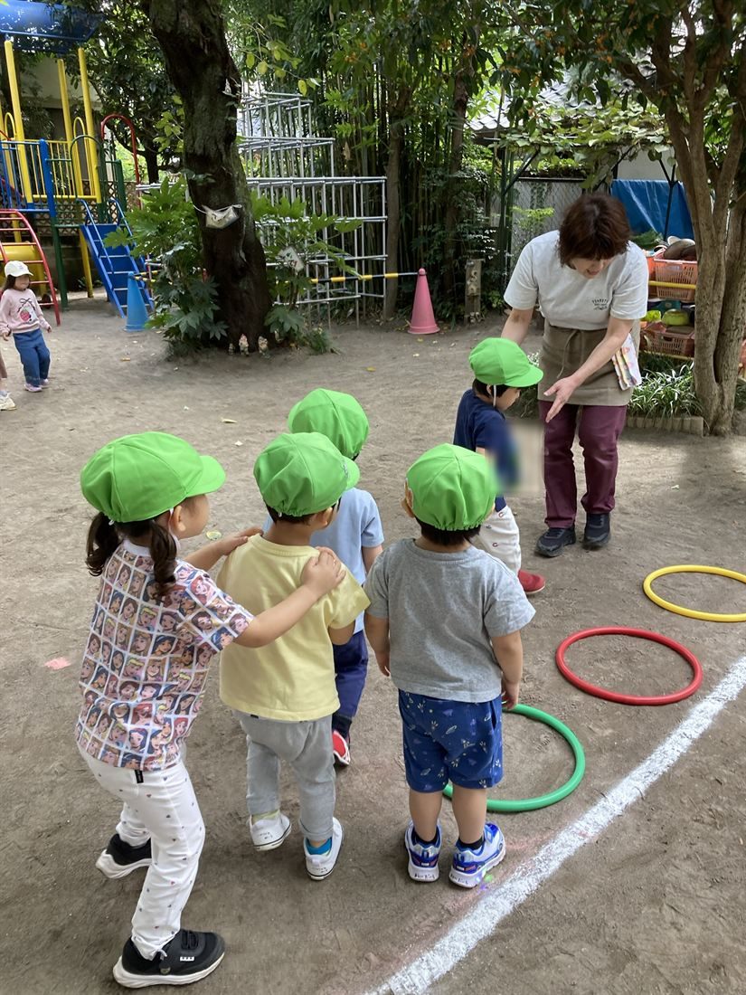 園庭で色とりどりのフープの前に並ぶ子どもたちと、腰をかがめた先生